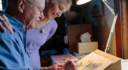 elderly woman watches her husband paint at his art desk