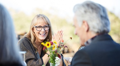 middle aged woman smiles at her date across outdoor dinner table with a vase of flowers
