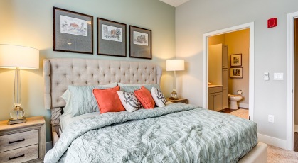 Model bedroom at our apartments Naperville, featuring wood grain floor paneling and a view of the bathroom. 