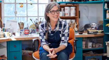 woman with glasses and overalls sits in her art studio, smiling
