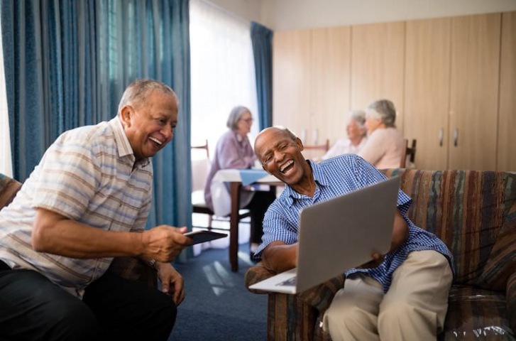 two-males-laughing-in-common-room-on-laptop-three-females-in-background-sitting-Overture